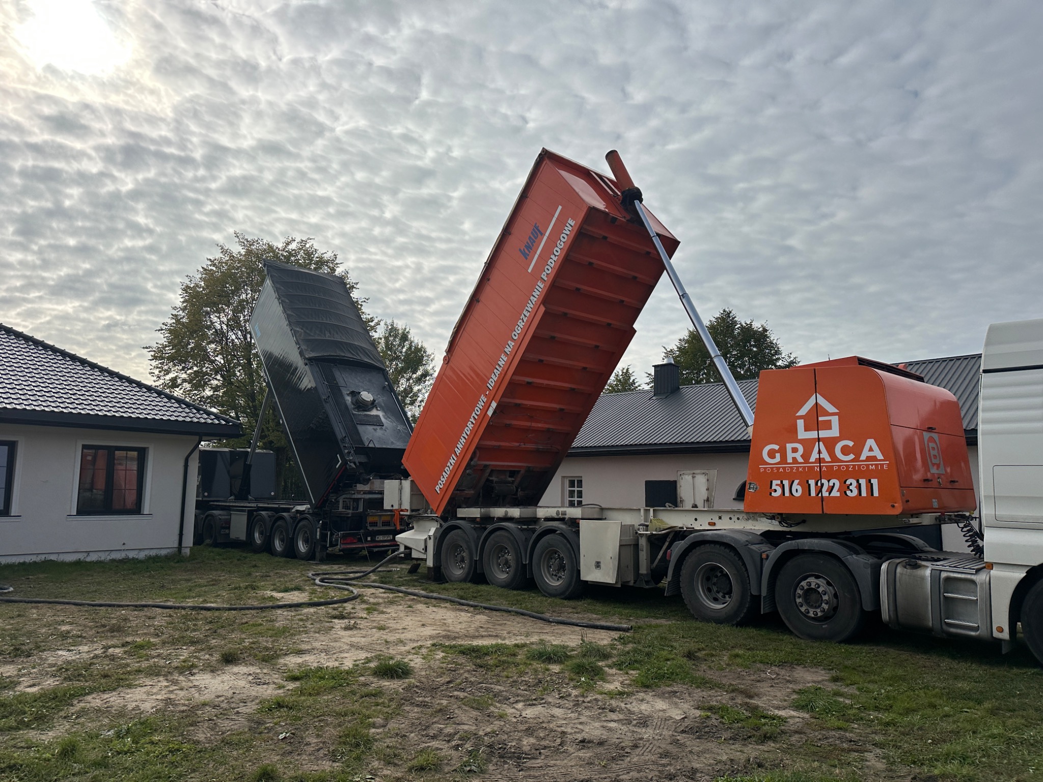 Dwa samochody ciężarowe z otwartymi naczepami, z których wylewana jest wylewka anhydrytowa. Widoczne węże i budynki w tle. Jeden z samochodów oznaczony logiem firmy GRACA.