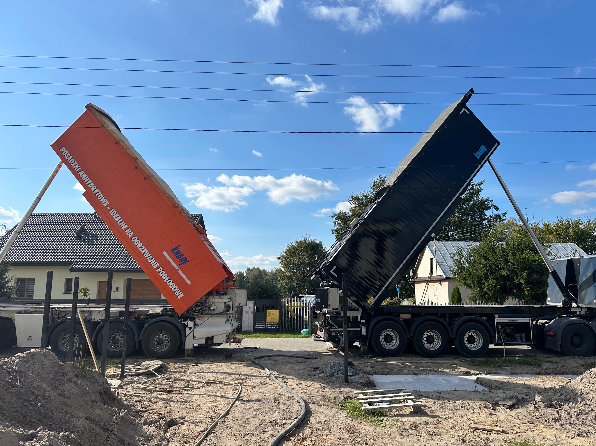 Dwa samochody ciężarowe z otwartymi skrzyniami, przygotowane do wylewania posadzki na budowie. Widoczne węże i teren przygotowany pod wylewkę. Błękitne niebo w tle.