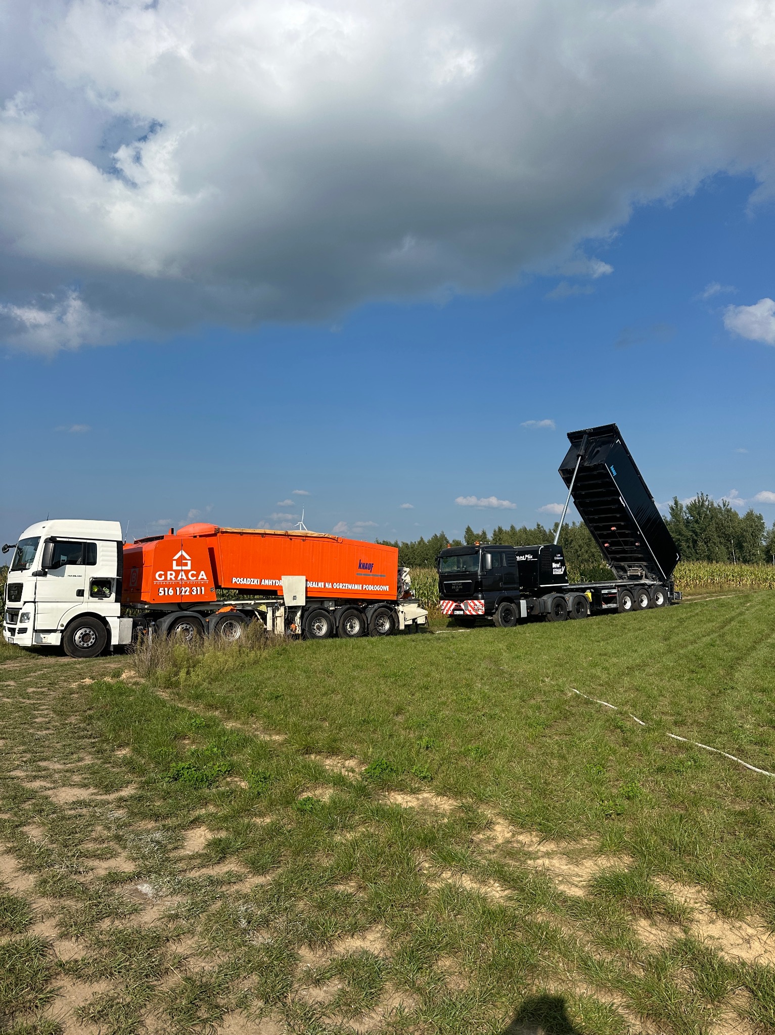 Dwa samochody ciężarowe: biały z pomarańczową cysterną Graca i czarny z podniesioną skrzynią ładunkową, na tle pola i błękitnego nieba z chmurami.