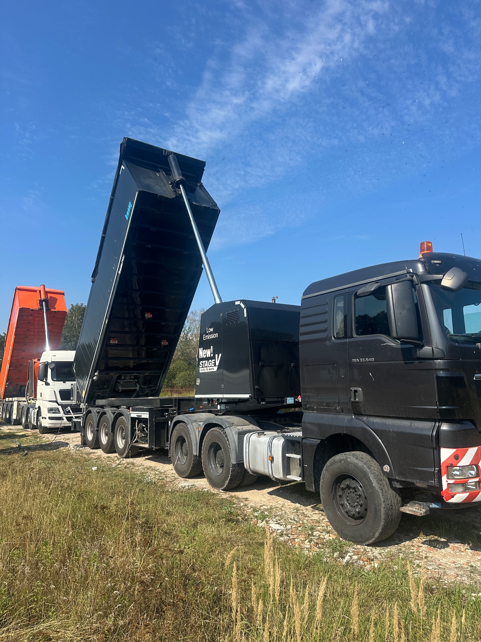 Ciężarówki z podniesionymi naczepami, prawdopodobnie do transportu materiałów sypkich na budowę. Widoczne detale techniczne pojazdów i otoczenie placu budowy.