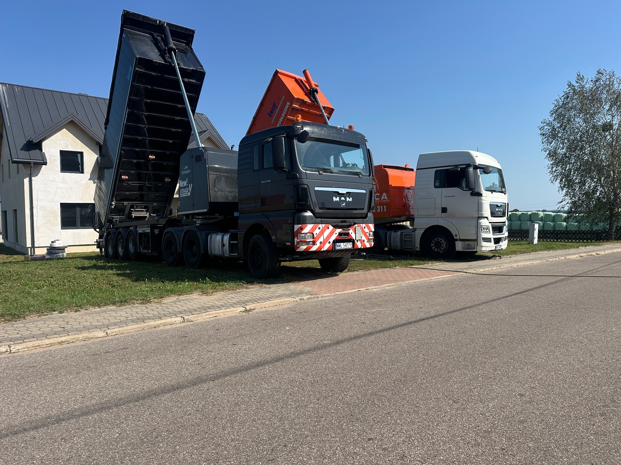 Ciężarówki MAN z podnośnikami do wylewek, zaparkowane obok domu. Na pomarańczowym podnośniku widoczny napis Knauf Podłogowe. Błękitne niebo w tle.