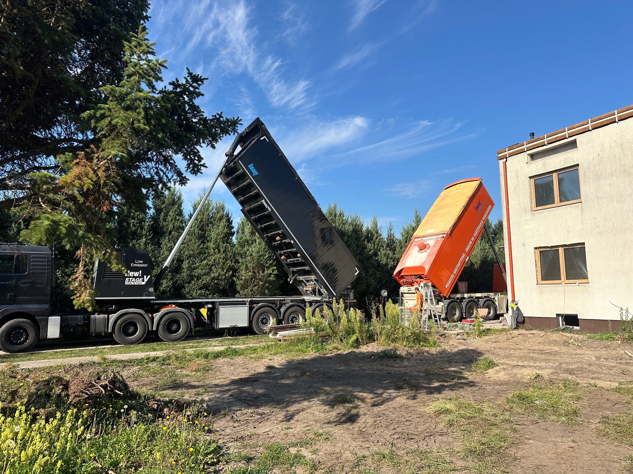 Dwa samochody ciężarowe z podniesionymi skrzyniami, jeden czarny, drugi pomarańczowy, na tle budynku i drzew. Widoczne prace przygotowawcze do wylewki.