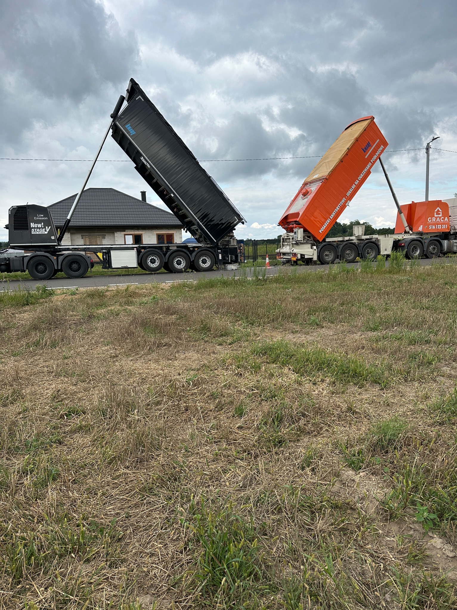 Dwa samochody ciężarowe z podniesionymi naczepami, jeden czarny z logo Knauf, drugi pomarańczowy z napisem 'Posadzki anhydrytowe', na tle domu i pochmurnego nieba.