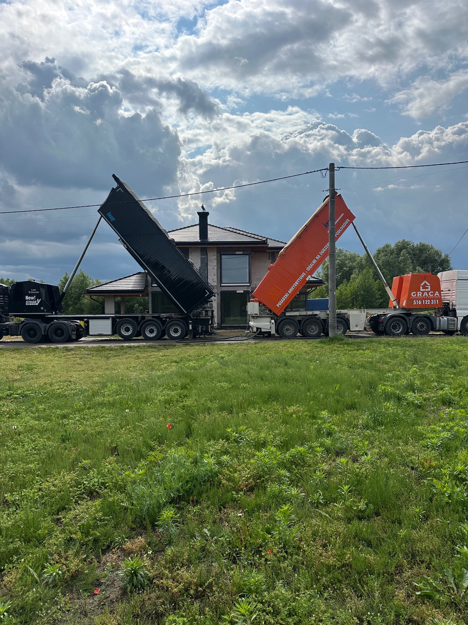 Dwa samochody ciężarowe z podniesionymi naczepami, jeden czarny z logo Knauf, drugi pomarańczowy z logo Graca, na tle domu w budowie i pochmurnego nieba.
