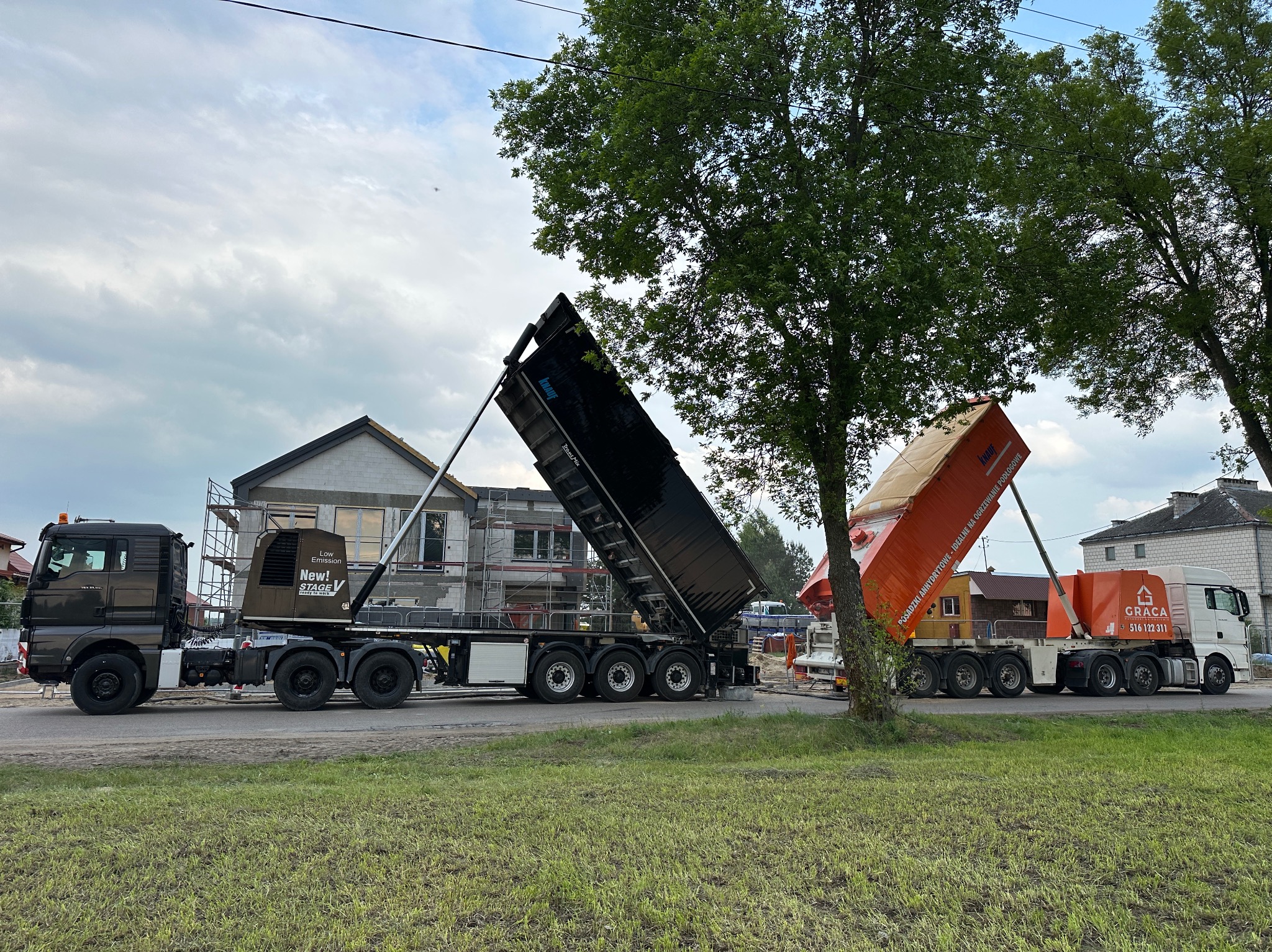 Dwa samochody ciężarowe z podniesionymi naczepami, prawdopodobnie podczas wylewania posadzki na budowie domu jednorodzinnego w tle. Jeden z nich ma logo firmy Graca.