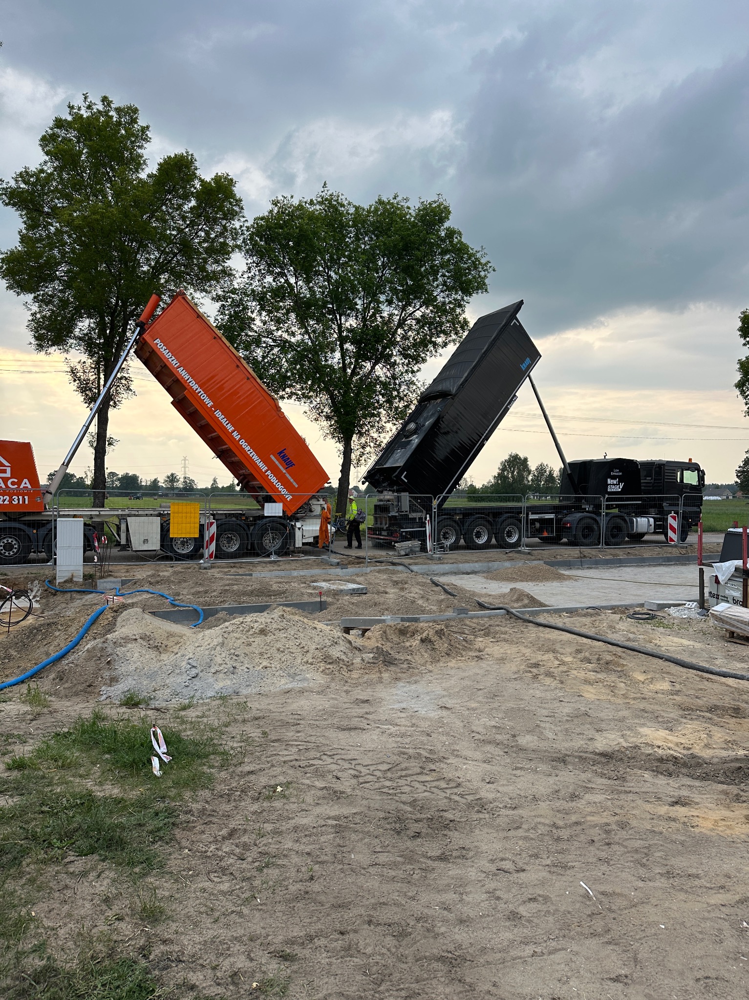 Dwa samochody ciężarowe z otwartymi skrzyniami, jeden pomarańczowy z napisem Knauf, drugi czarny, przygotowują się do wylania materiału na plac budowy pod posadzki.