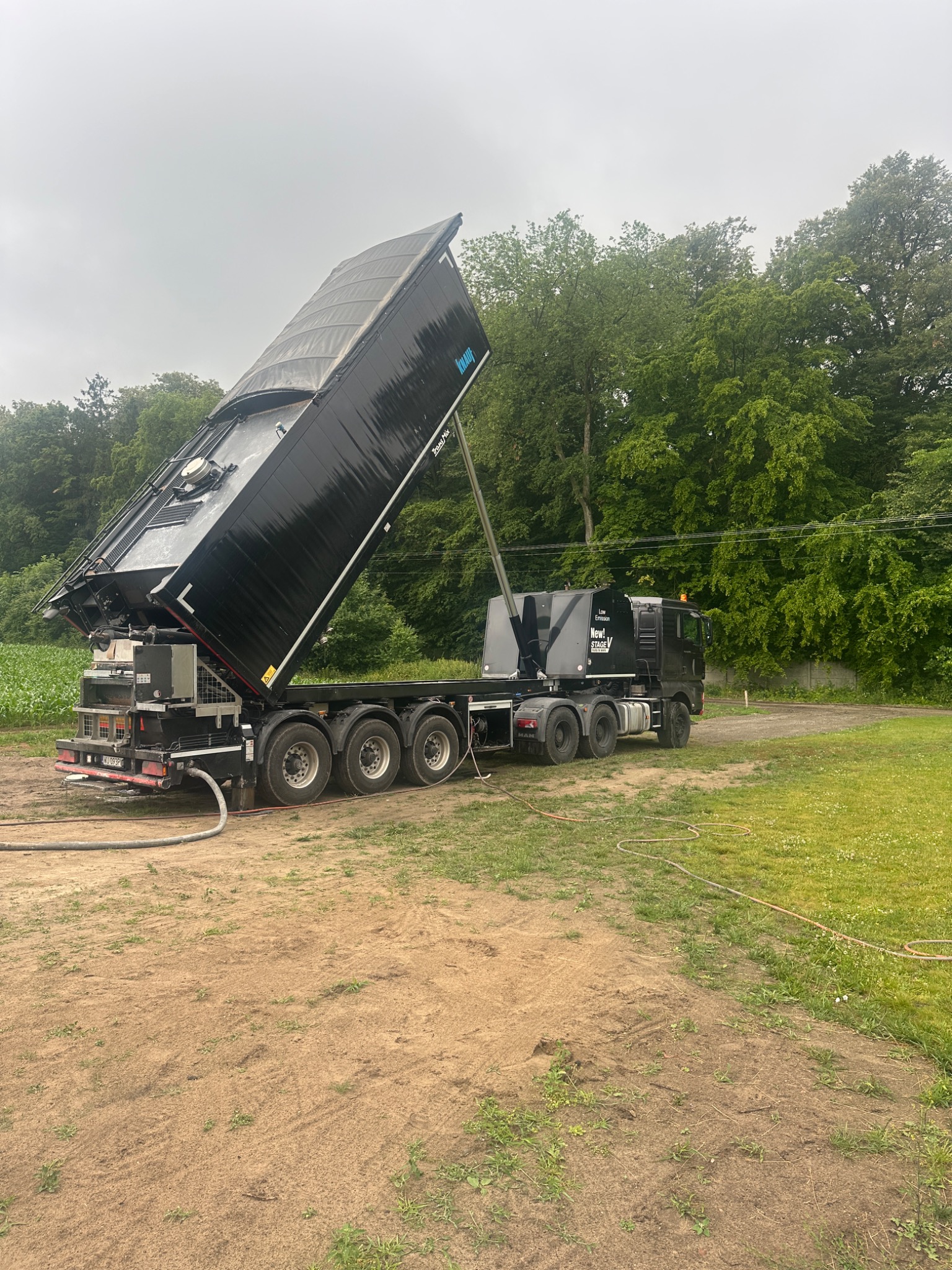 Czarny samochód ciężarowy z podniesioną naczepą na tle zieleni, prawdopodobnie przygotowany do wylewania mieszanki. Widoczny wąż i teren przygotowany pod wylewkę.