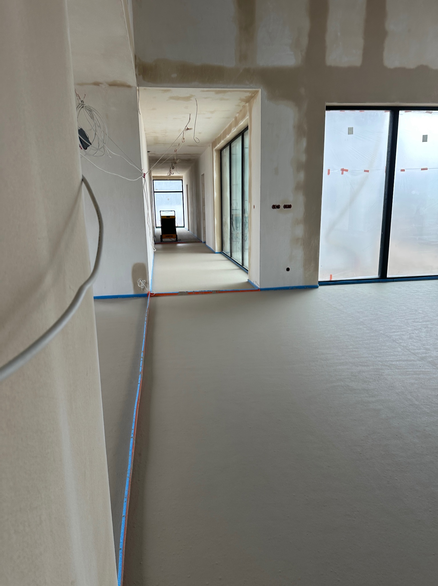 Gładka, szara posadzka w długim korytarzu z dużymi oknami, zabezpieczona niebieską taśmą. Widoczne kable i surowe ściany w trakcie wykończenia.