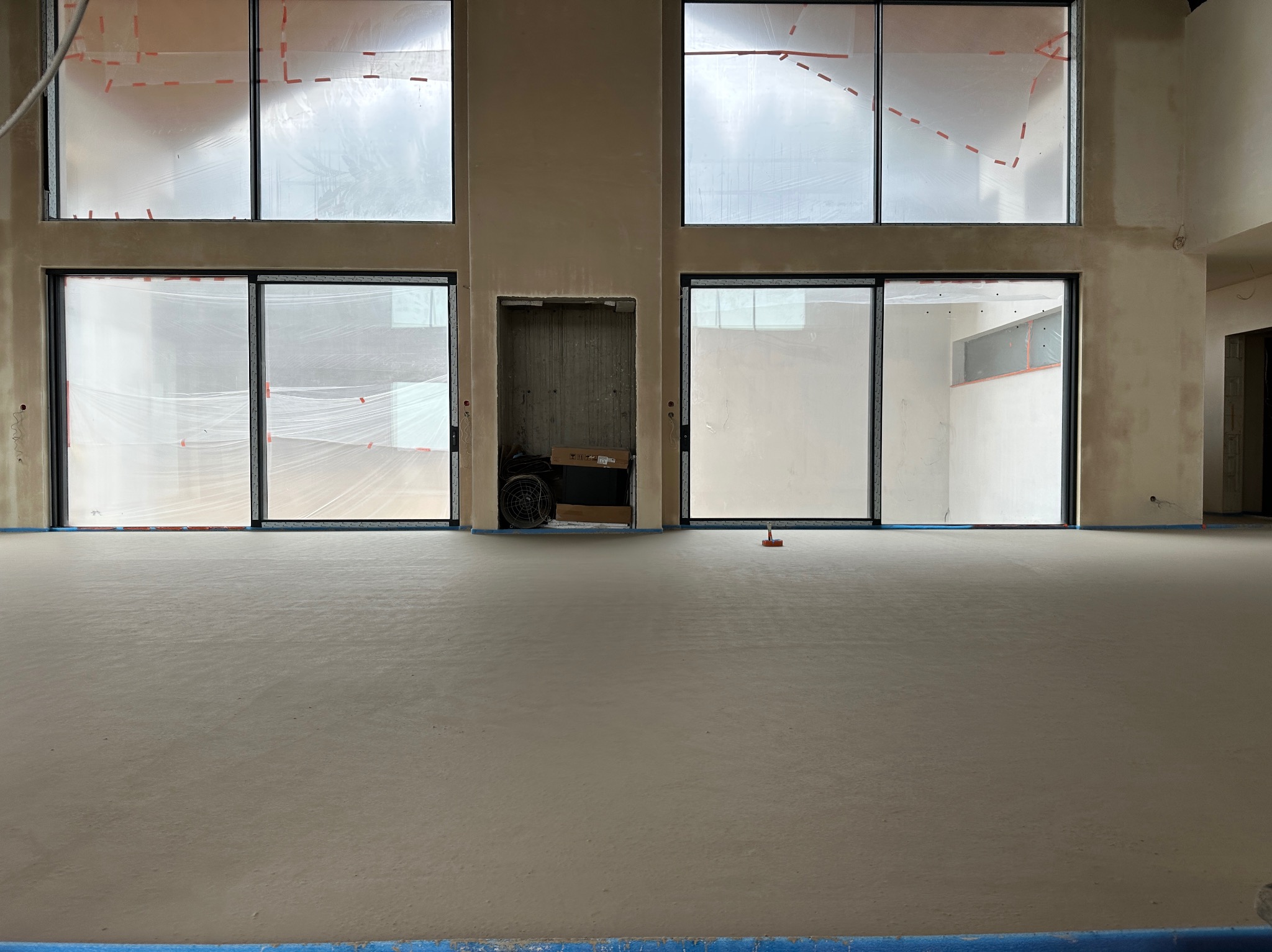 Jasna, gładka posadzka w przestronnym wnętrzu z dużymi oknami zabezpieczonymi folią. Widoczny fragment wentylatora i narzędzie do rozprowadzania masy.