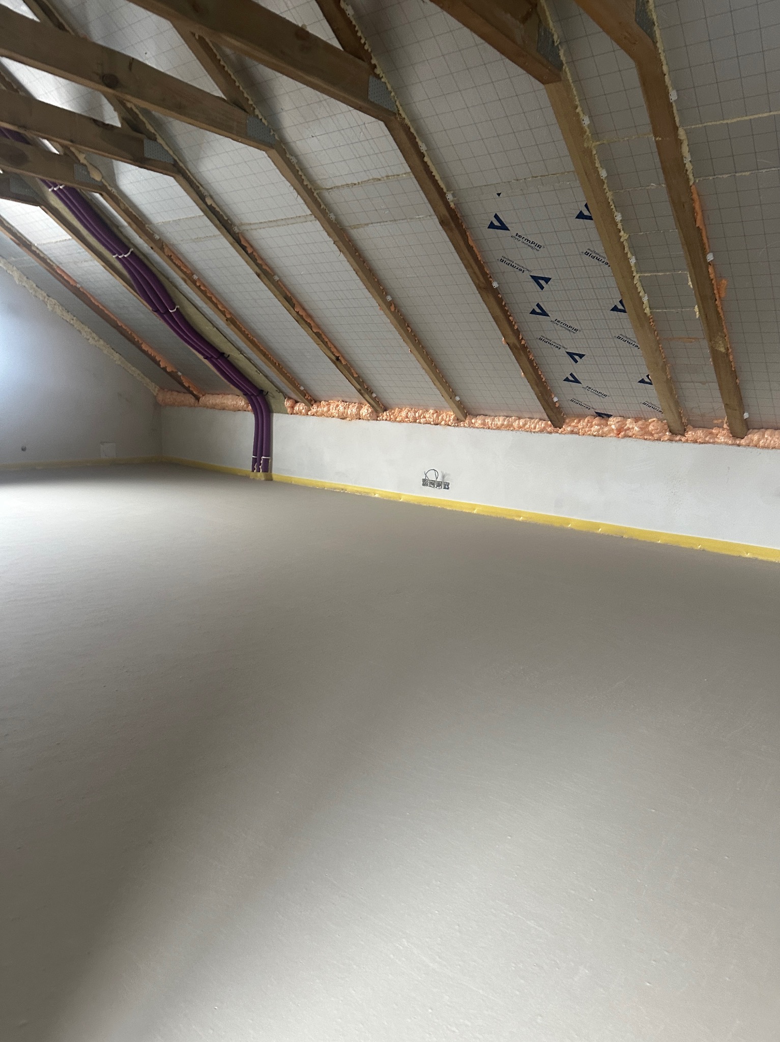 Gładka, szara posadzka w nowym poddaszu z widoczną izolacją stropu i fioletowymi rurami przy ścianie. Żółta taśma dylatacyjna wzdłuż ściany.