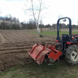 Czerwony traktor z glebogryzarką na polu, świeżo przygotowana ziemia w rzędach, w tle kwitnące drzewa i brzoza.