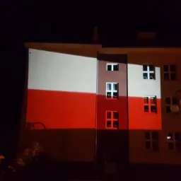 Budynek oświetlony w barwach flagi Polski nocą, widoczne okna i lampa uliczna.