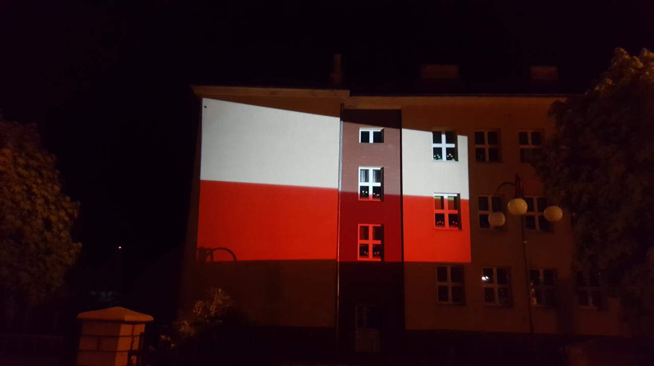 Budynek oświetlony w barwach flagi Polski nocą, widoczne okna i lampa uliczna.