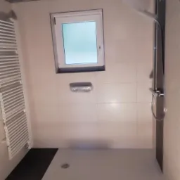 Niedokończona łazienka z jasnymi płytkami, brodzikiem z odpływem liniowym, grzejnikiem drabinkowym i czarną armaturą prysznicową; surowy strop z widoczną konstrukcją.