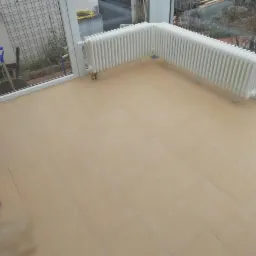 Jasne, podłużne płytki ułożone na podłodze w oranżerii, z grzejnikiem przy ścianie i widokiem na ogród przez okno.