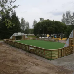 Nowo wybudowane boisko sportowe z drewnianym ogrodzeniem i sztuczną trawą, otoczone ziemią i drzewami.