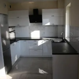 Nowoczesna biała kuchnia z czarnym blatem i płytkami w geometryczne wzory, oświetlona naturalnym światłem z okna.