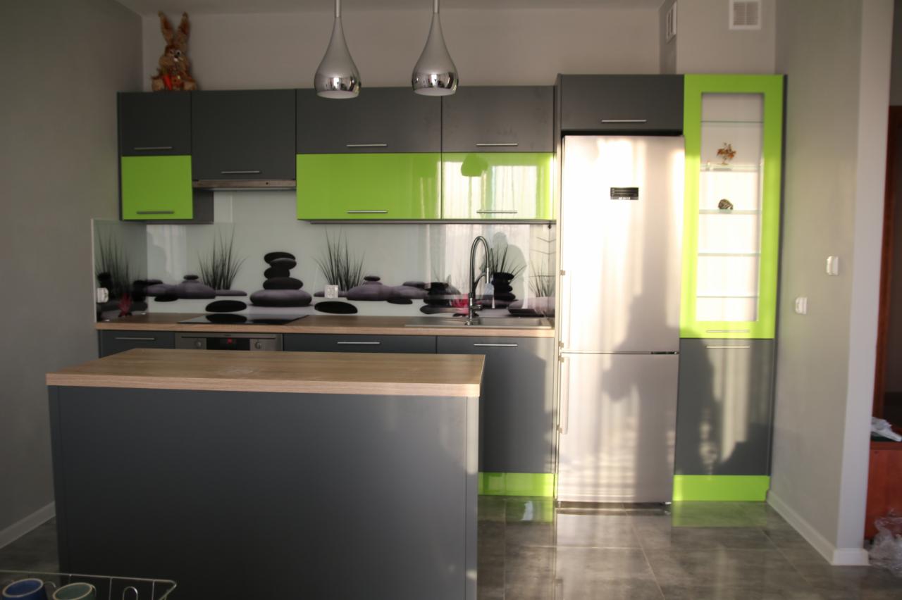 Nowoczesna kuchnia z szarymi szafkami, zielonymi akcentami i wyspą kuchenną z drewnianym blatem. Na ścianie szklany panel z motywem kamieni zen.