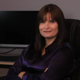 Portret uśmiechniętej kobiety w fioletowej satynowej bluzce siedzącej na czarnym skórzanym krześle biurowym z podłokietnikami, na tle biurka z monitorem.