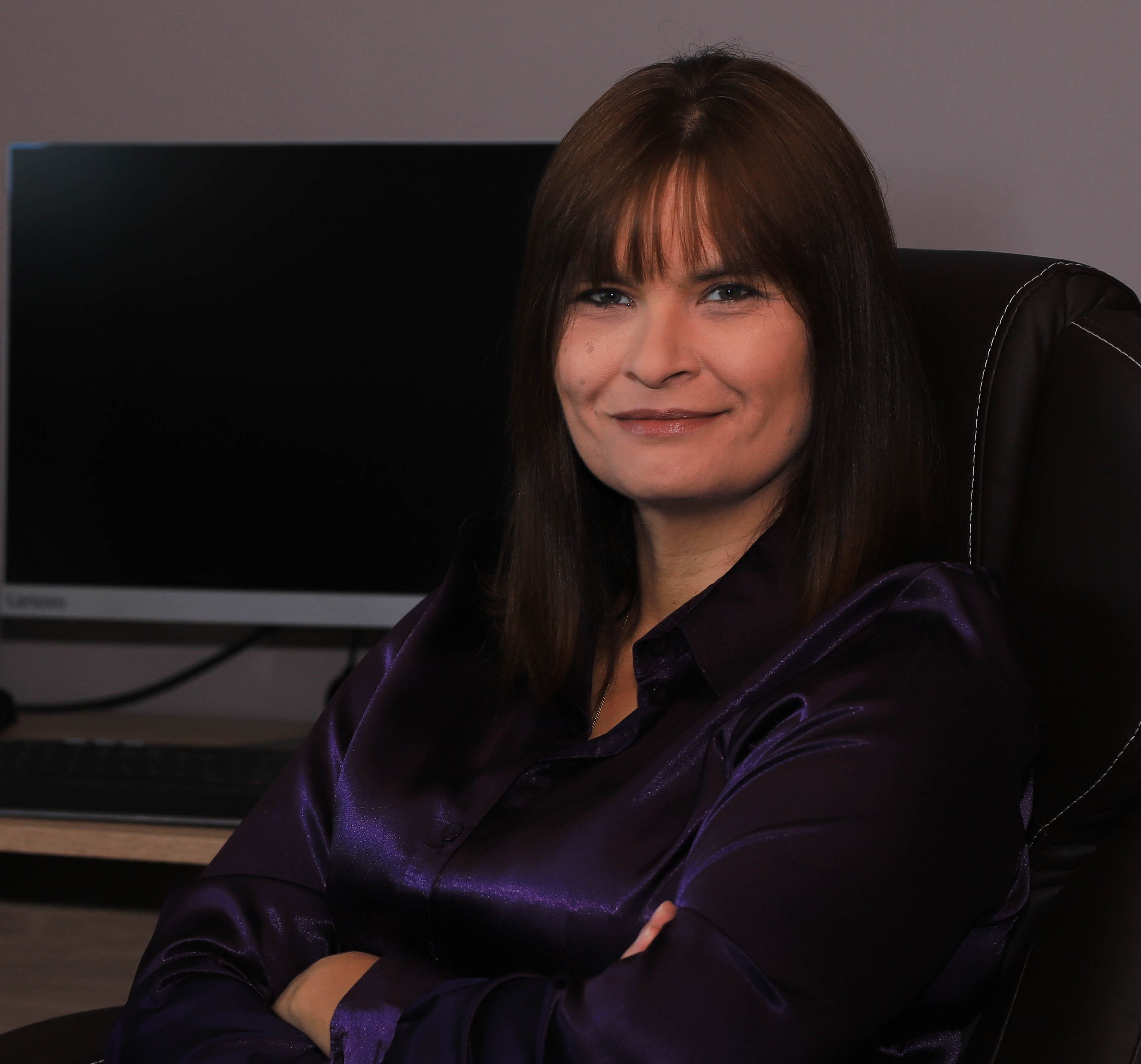 Portret uśmiechniętej kobiety w fioletowej satynowej bluzce siedzącej na czarnym skórzanym krześle biurowym z podłokietnikami, na tle biurka z monitorem.