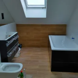 Wnętrze łazienki w trakcie wykańczania, z widoczną białą wanną obudowaną czarnymi panelami, wiszącą miską WC, umywalką z szafką zabezpieczoną folią, brązowymi płytkami imitującymi drewno...