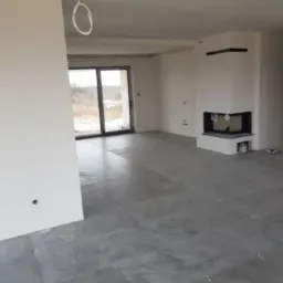 Szare płytki podłogowe ułożone w przestronnym, jasnym salonie z kominkiem; widoczne krzyżyki dystansowe między płytkami, surowy stan wykończenia.