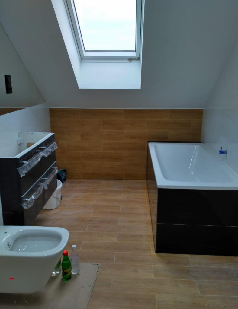 Wnętrze łazienki w trakcie wykańczania, z widoczną białą wanną obudowaną czarnymi panelami, wiszącą miską WC, umywalką z szafką zabezpieczoną folią, brązowymi płytkami imitującymi drewno...