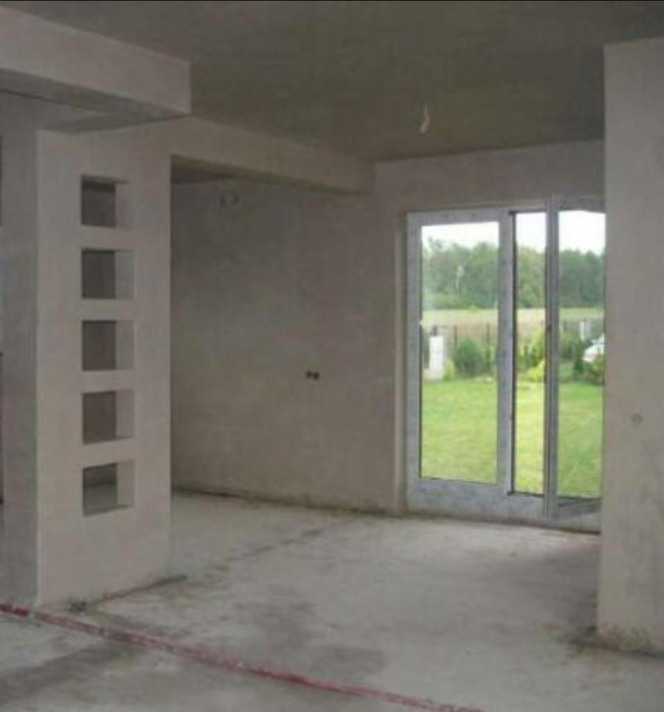 Surowy stan wnętrza: betonowa podłoga, ściany w szarym tynku, kolumna z kwadratowymi wnękami i przeszklone drzwi przesuwne z widokiem na zieleń.