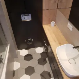 Nowoczesna łazienka z heksagonalną podłogą w odcieniach szarości, białą umywalką nablatową i drewnianym blatem, widoczne przygotowane otwory na armaturę podtynkową.