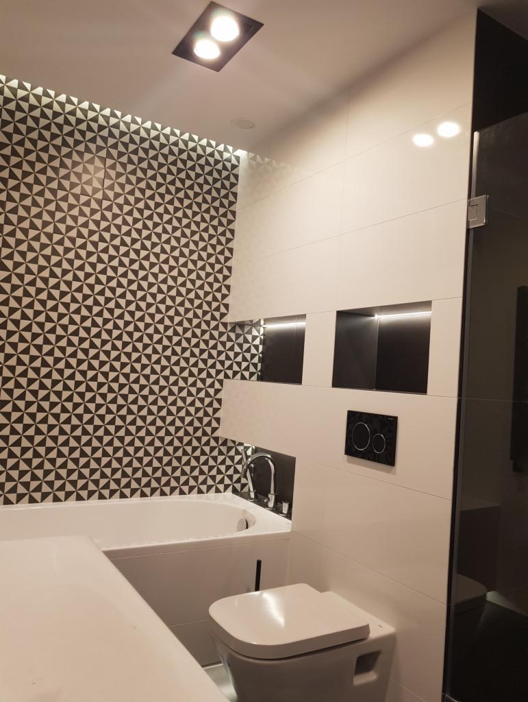 Nowoczesna łazienka z czarno-białą mozaiką na ścianie, białą wanną, toaletą i podtynkową baterią umywalkową, oświetlona dwoma punktowymi lampami sufitowymi.