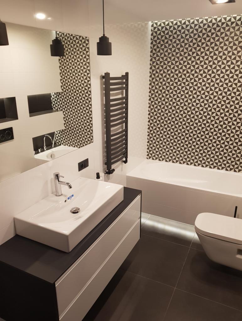 Nowoczesna łazienka z białą prostokątną umywalką nablatową, czarnym blatem i szafką z białymi frontami, grzejnikiem drabinkowym w kolorze grafitowym, białą wanną oraz toaletą. Ściany wyłożone...