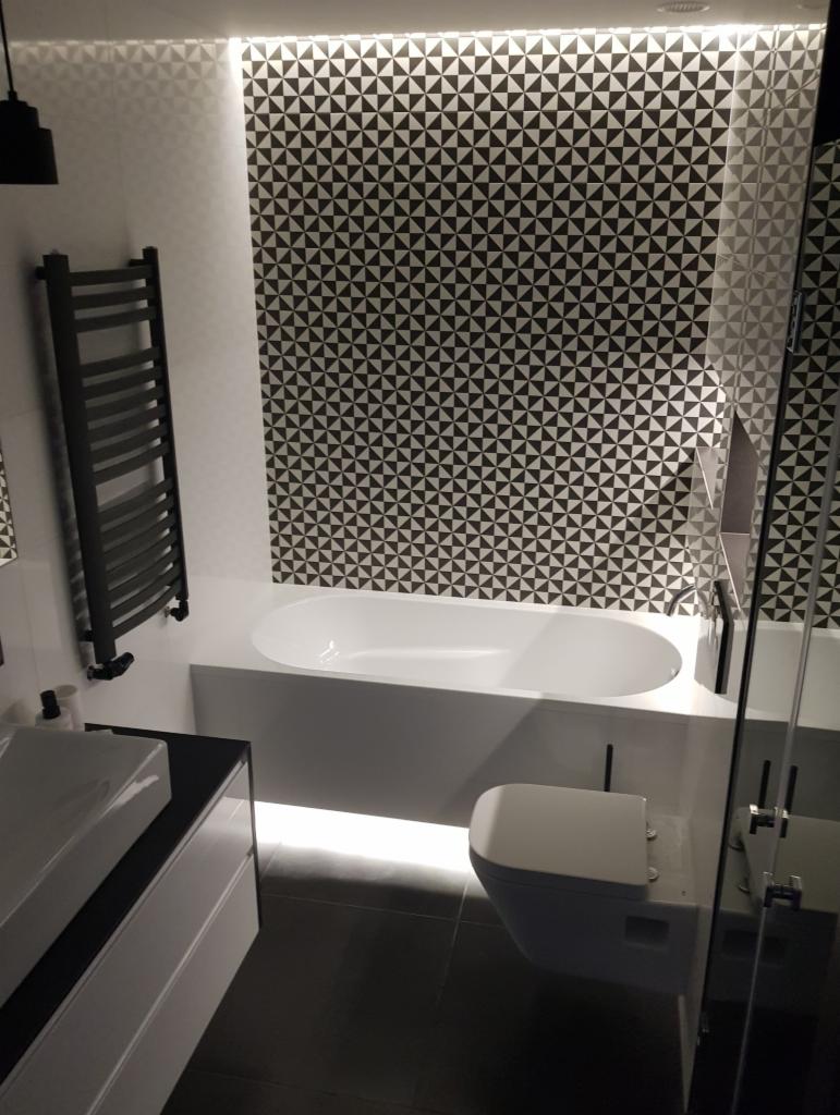 Nowoczesna łazienka w czarno-białej kolorystyce z geometrycznym wzorem na ścianie, podświetlaną wanną i wiszącą toaletą.