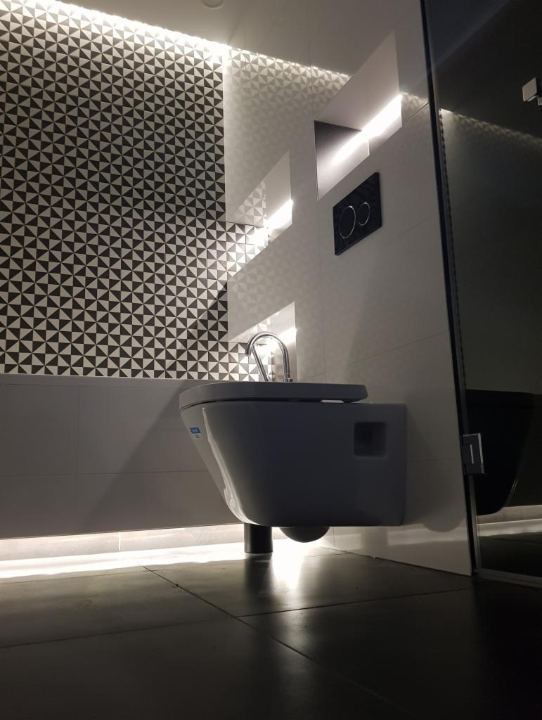 Nowoczesna toaleta podwieszana z chromowaną armaturą na tle ściany wyłożonej biało-czarną mozaiką i podświetlonej listwą LED, widok z niskiego kąta.