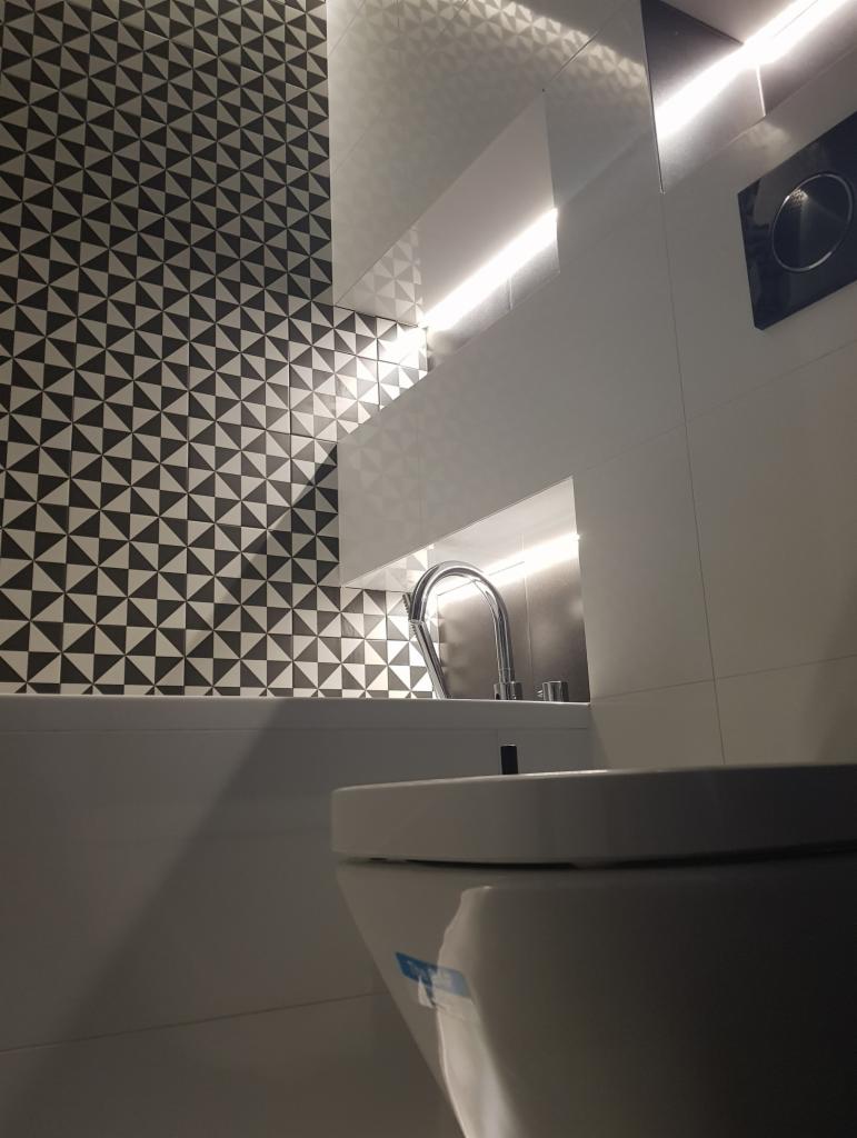 Nowoczesna łazienka z geometryczną mozaiką na ścianie, podświetlanym lustrem i chromowaną armaturą, widok na toaletę z bliska.
