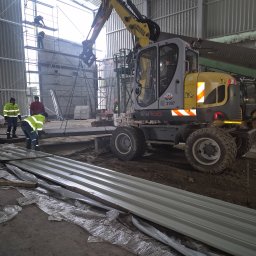 PSJ JANUSZ STANIK - Żółto-szara koparka Wacker Neuson EW100 podnosi metalowy element na placu budowy w hali, robotnicy w odblaskowych kamizelkach w tle, widoczne rusztowania i materiały budowlane.