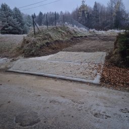PSJ JANUSZ STANIK - Betonowy przepust z kratką odwadniającą na drodze gruntowej w zimowej scenerii, drzewa i trawa pokryte szronem.