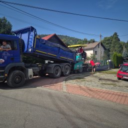 PSJ JANUSZ STANIK - Niebieska ciężarówka wywrotka MAN TGA 26.350 z logiem firmy PSJ rozładowuje materiał do rozściełacza Vögele Super 1303-3 podczas prac drogowych na tle domów i zieleni, widoczny pracownik...