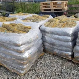 Palety z workami wypełnionymi ziemią ogrodową, owinięte folią stretch, na żwirowym podłożu, w tle widoczne ogrodzenie i roślinność.