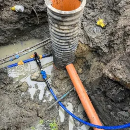 Wykop z widocznym pionowym, żebrowanym elementem studzienki kanalizacyjnej, pomarańczową rurą kanalizacyjną pod kątem, niebieskim przewodem i mokrą ziemią.