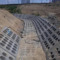 Ułożone betonowe ażurowe płyty koryta odwadniającego, w tle zbiornik przemysłowy i ogrodzenie z siatki.