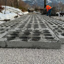 PSJ JANUSZ STANIK - Układanie betonowych płyt ażurowych na podsypce żwirowej w górskim krajobrazie; pracownik w pomarańczowej kurtce wyrównuje teren, a w tle widoczny samochód dostawczy i zalesione wzgórza o zmierzchu.