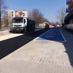 Układanie asfaltu na nowym parkingu z wyznaczonymi miejscami dla niepełnosprawnych, widoczny walec drogowy w oddali i ciężarówka z tyłu.