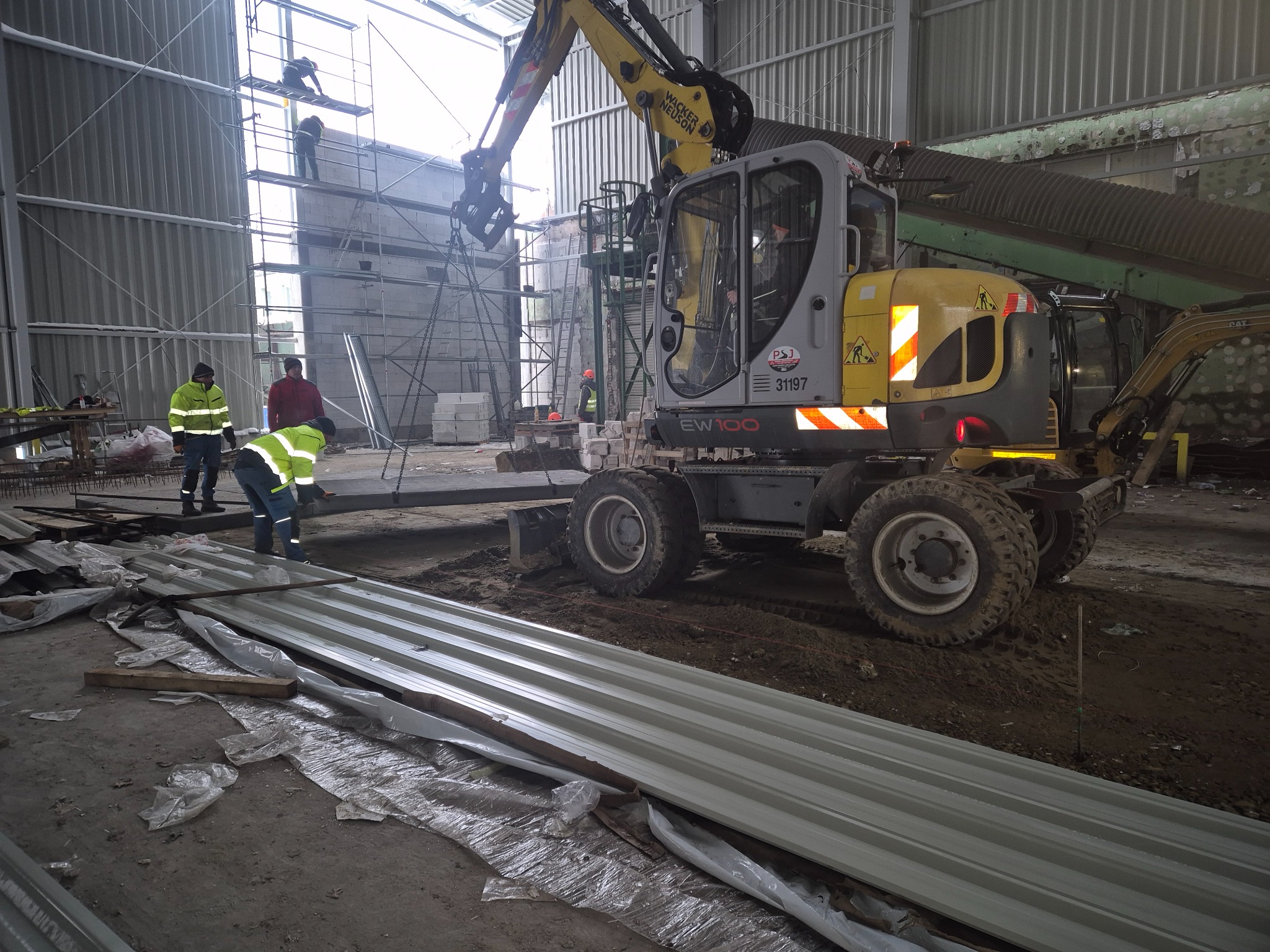 Żółto-szara koparka Wacker Neuson EW100 podnosi metalowy element na placu budowy w hali, robotnicy w odblaskowych kamizelkach w tle, widoczne rusztowania i materiały budowlane.