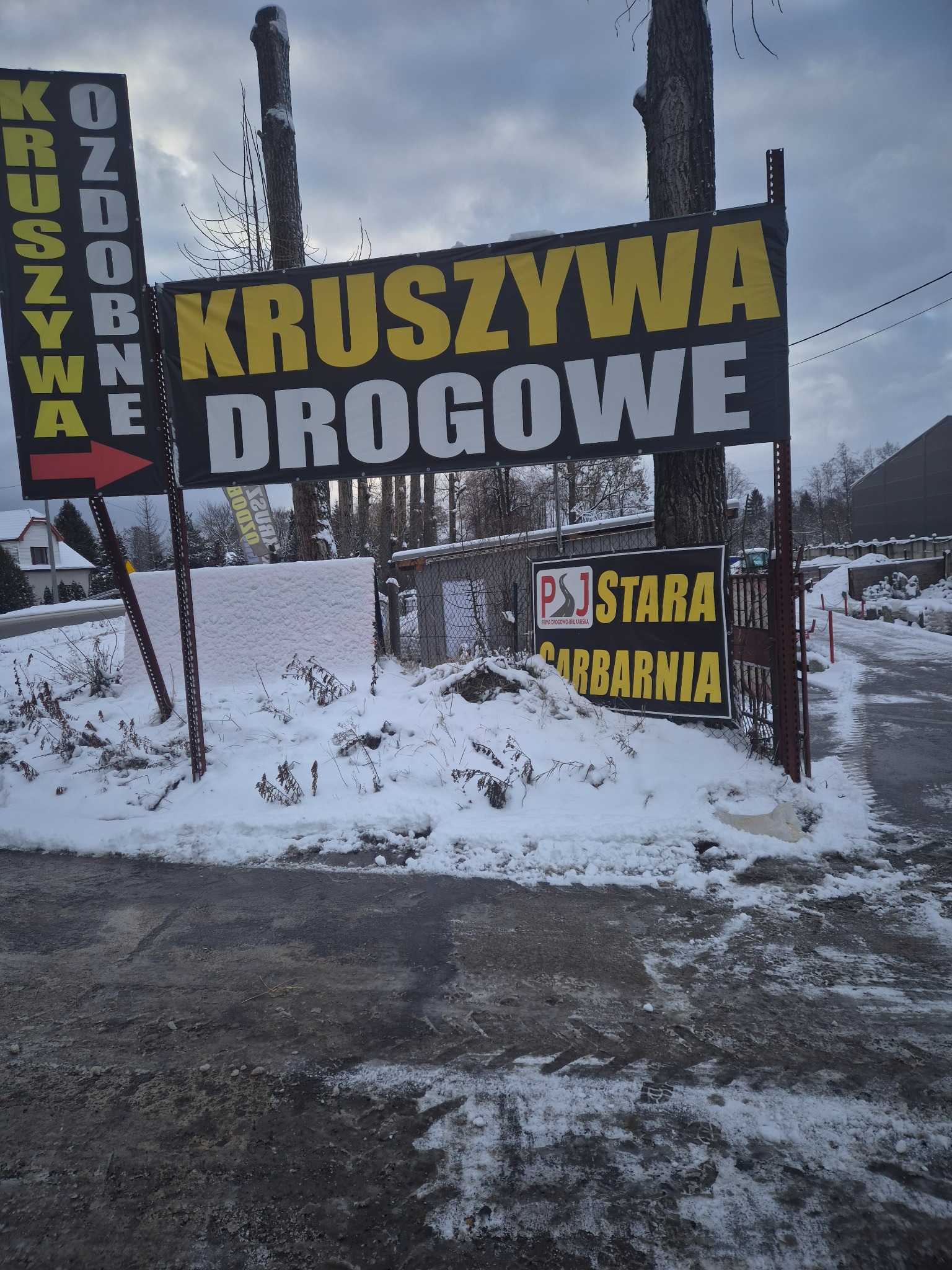 Zimowy widok na tablice reklamowe firmy PSJ Stara Garbarnia oferującej kruszywa drogowe i ozdobne, ustawione przy zasypanej śniegiem drodze.