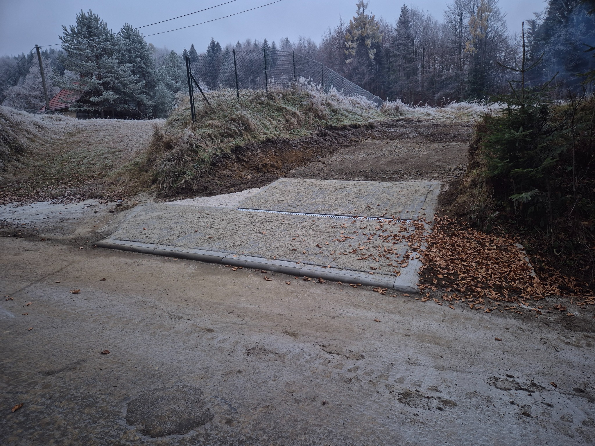 Betonowy przepust z kratką odwadniającą na drodze gruntowej w zimowej scenerii, drzewa i trawa pokryte szronem.