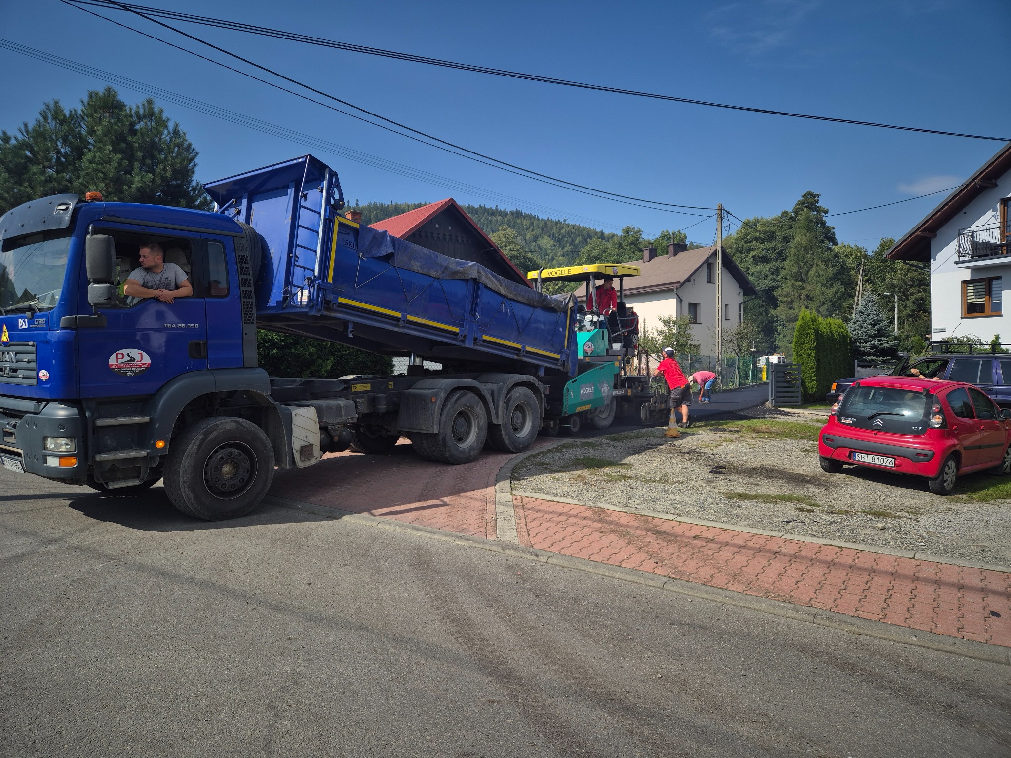 Niebieska ciężarówka wywrotka MAN TGA 26.350 z logiem firmy PSJ rozładowuje materiał do rozściełacza Vögele Super 1303-3 podczas prac drogowych na tle domów i zieleni, widoczny pracownik...