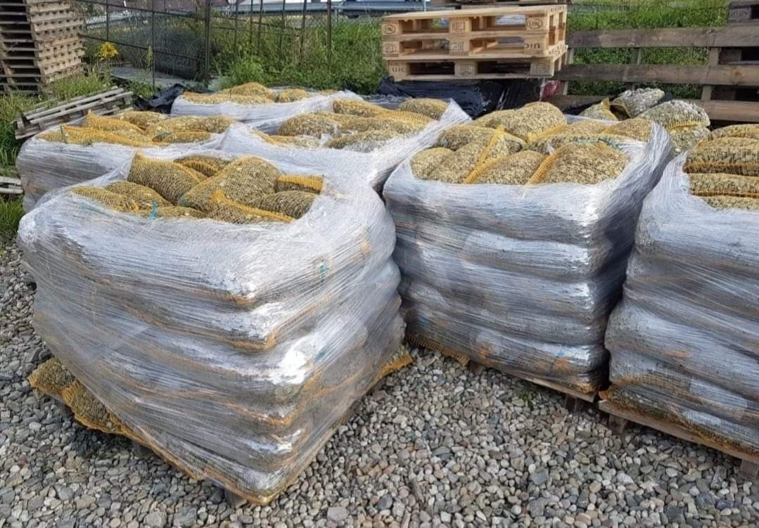 Palety z workami wypełnionymi ziemią ogrodową, owinięte folią stretch, na żwirowym podłożu, w tle widoczne ogrodzenie i roślinność.