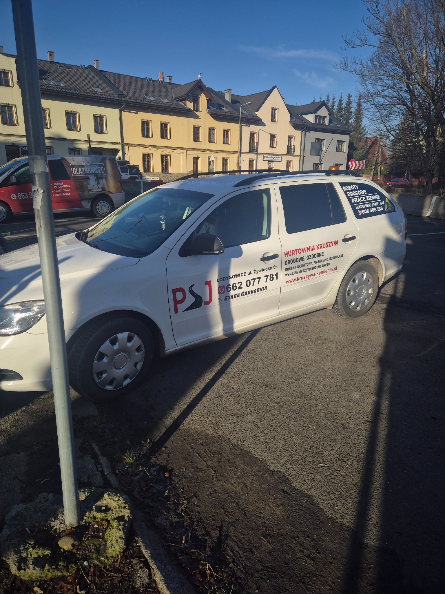 Biała Skoda Octavia kombi z naklejkami reklamowymi firmy Hurtownia Kruszyw i PSJ, zaparkowana na ulicy w słoneczny dzień, w tle budynki mieszkalne.