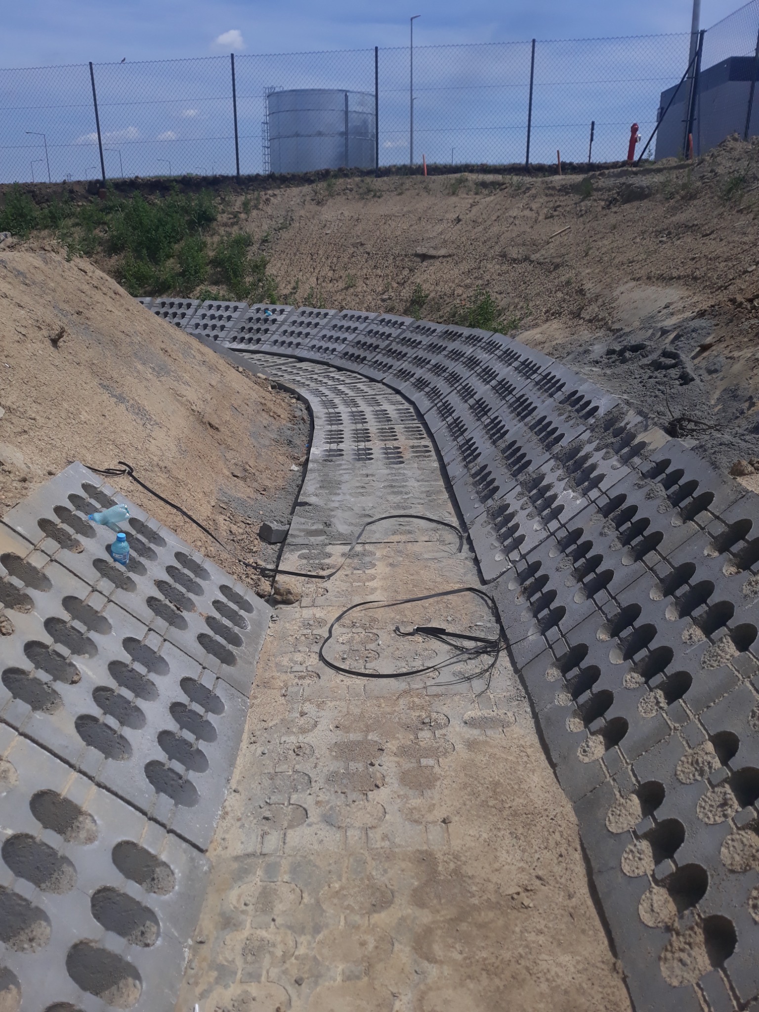 Ułożone betonowe ażurowe płyty koryta odwadniającego, w tle zbiornik przemysłowy i ogrodzenie z siatki.