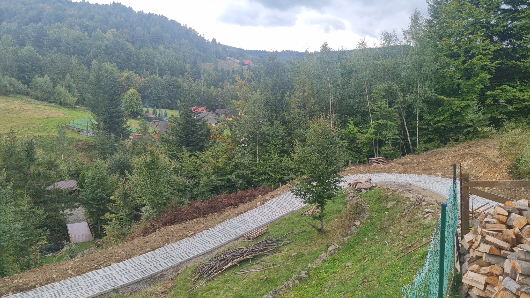 Ułożona z ażurowych płyt betonowych kręta droga na zboczu, w tle las i zabudowania, obok stos drewna i zielona siatka ogrodzeniowa.